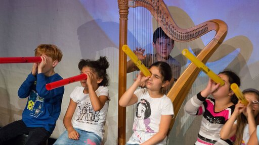 Viele Kinder schauen durch ein Plastikrohr, dahinter steht ein Junge mit einer Harfe.