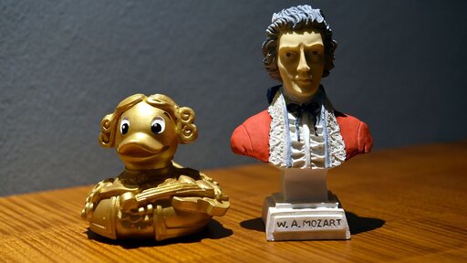 Zwei kleine Figuren, die linke goldene ist eine Quietscheente, die aussieht wie Mozarte, die rechte ist eine Büste von Mozart.